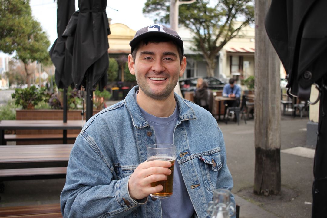 Hayden smiling in a denim jacket and cap, holding a beer at an outdoor pub table